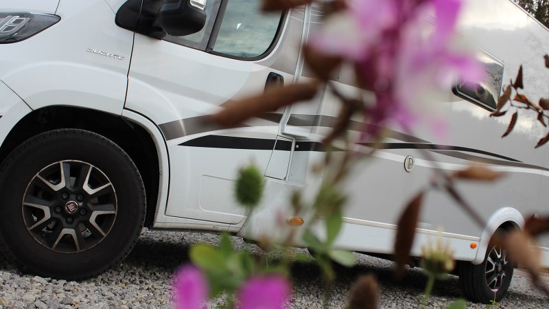 Weißes Wohnmobil auf Kiesplatz, im Vordergrund unscharfe Blumen. Seitliche Ansicht mit Fokus auf die Räder., © Erlebnisregion Schwäbischer Albtrauf e. V. Weißes Wohnmobil auf Kiesplatz, im Vordergrund unscharfe Blumen. Seitliche Ansicht mit Fokus auf die Räder., © Erlebnisregion Schwäbischer Albtrauf e. V.