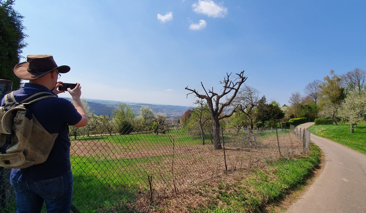 Person mit Hut fotografiert eine Landschaft mit Streuobstwiesen. Ein Weg führt durch die grüne Landschaft, der Himmel ist blau mit wenigen Wolken., © Kulturamt Plochingen Person mit Hut fotografiert eine Landschaft mit Streuobstwiesen. Ein Weg führt durch die grüne Landschaft, der Himmel ist blau mit wenigen Wolken., © Kulturamt Plochingen