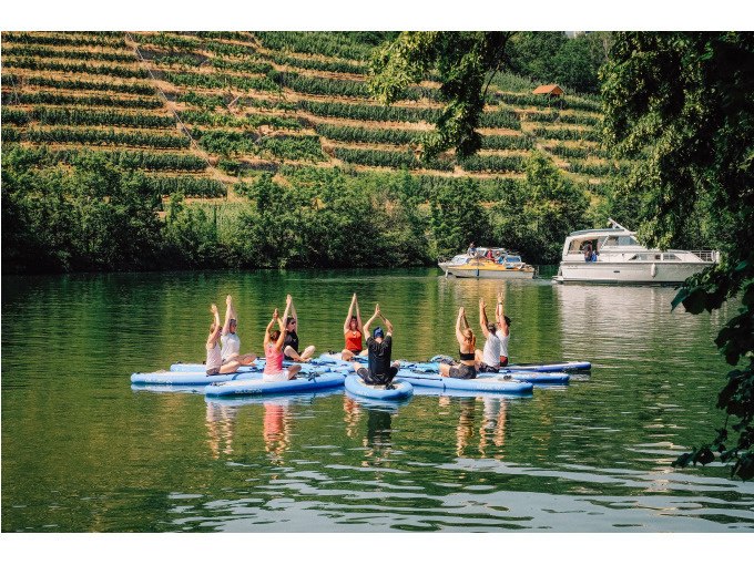 Menschen praktizieren Yoga auf SUP-Boards auf einem Fluss, umgeben von Weinbergen und einem vorbeifahrenden Boot., © Cool-Tours StattReisen Menschen praktizieren Yoga auf SUP-Boards auf einem Fluss, umgeben von Weinbergen und einem vorbeifahrenden Boot., © Cool-Tours StattReisen