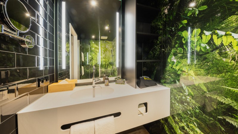 Modernes Badezimmer mit wei&szlig;em Waschbecken, Spiegel und Dschungelmotiv an der Wand. Schwarze Fliesen und helle Beleuchtung erg&auml;nzen das Design., &copy; Rioca Stuttgart Posto 4 GmbH