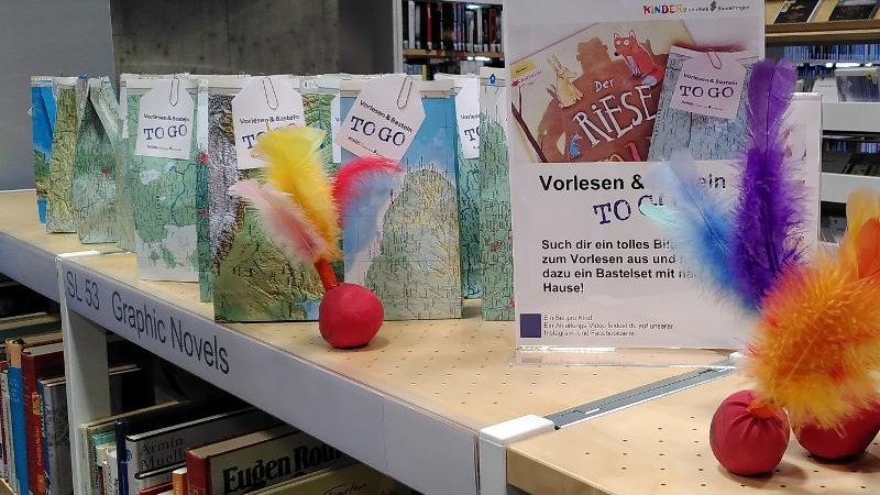 Regal in Bibliothek mit Bastelsets und Schild 'Vorlesen & Basteln to go'. Bunte Federn und Bücher im Hintergrund., © Stadtbibliothek Sindelfingen