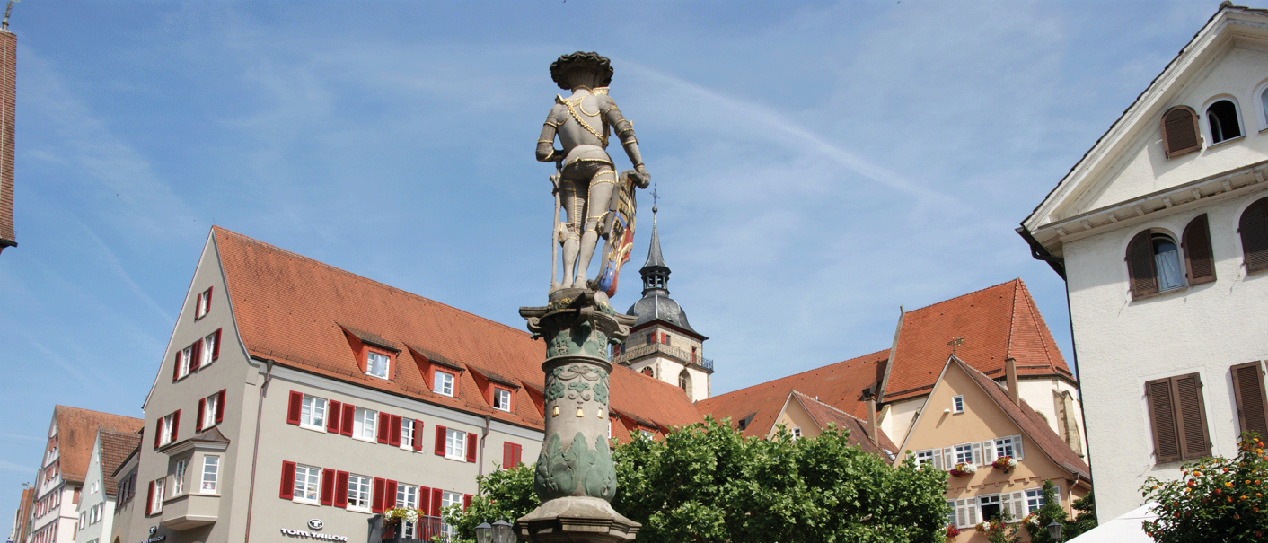 Marktbrunnen mit Statue auf einem belebten Platz, umgeben von historischen Gebäuden mit roten Dächern und Bäumen., © Stuttgart-Marketing GmbH