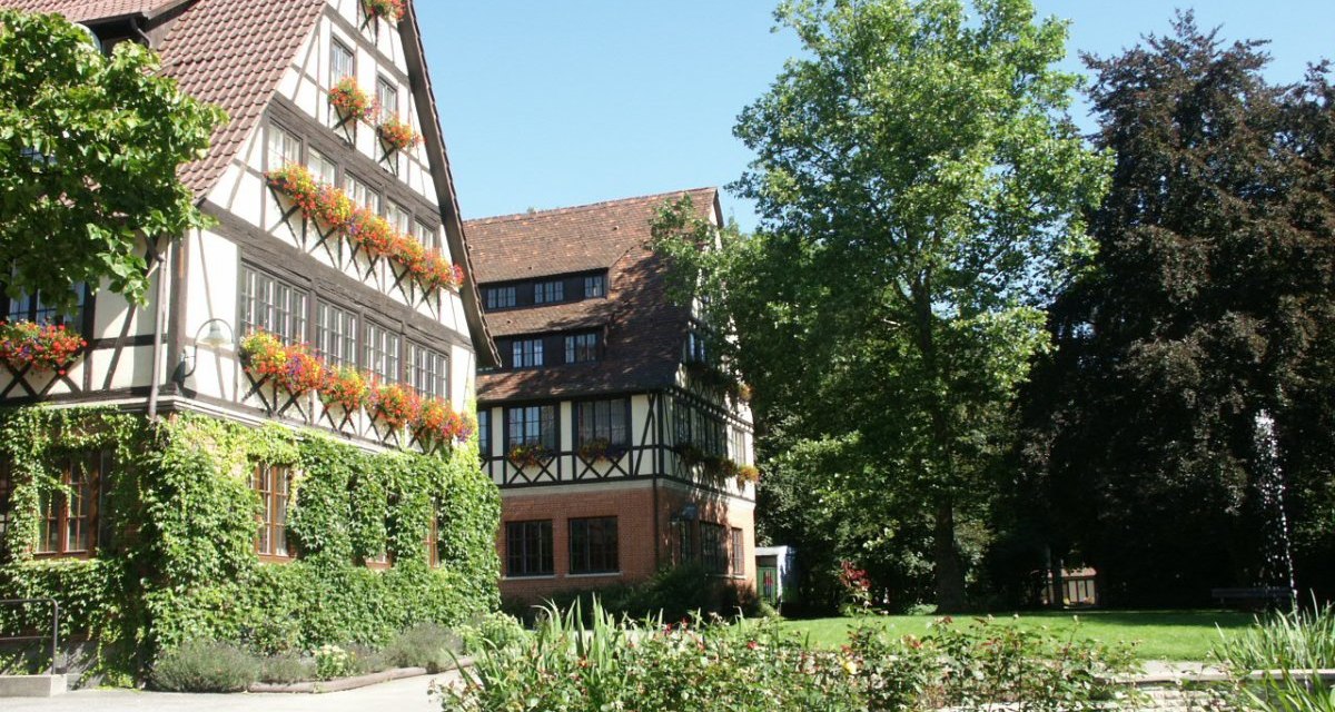 Fachwerkhaus mit blühenden Blumenkästen und grünem Efeu. Im Vordergrund ein gepflegter Garten mit Brunnen, umgeben von Bäumen., © Natur.Nah. Schönbuch & Heckengäu Fachwerkhaus mit blühenden Blumenkästen und grünem Efeu. Im Vordergrund ein gepflegter Garten mit Brunnen, umgeben von Bäumen., © Natur.Nah. Schönbuch & Heckengäu