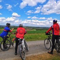 Drei Radfahrer in bunter Sportkleidung stehen auf einem Feldweg und schauen in die weite, grüne Landschaft unter einem blauen Himmel mit Wolken., © Land der 1000 Hügel - Kraichgau-Stromberg Drei Radfahrer in bunter Sportkleidung stehen auf einem Feldweg und schauen in die weite, grüne Landschaft unter einem blauen Himmel mit Wolken., © Land der 1000 Hügel - Kraichgau-Stromberg
