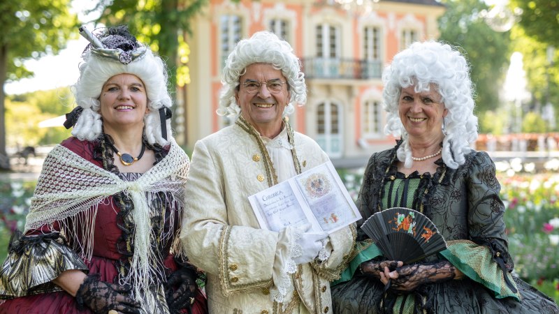 Drei Personen in barocken Kostümen posieren lächelnd vor einem historischen Gebäude. Sie tragen Perücken und halten Accessoires wie einen Fächer und ein Buch., © Für die Bildrechte: Amt für Medien und Kommunikation Schwäbisch Gmünd