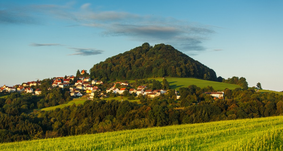 Der Berg Hohenstaufen erhebt sich hinter einem Dorf, umgeben von grünen Feldern und Bäumen, unter einem klaren blauen Himmel., © Landkreis Göppingen Der Berg Hohenstaufen erhebt sich hinter einem Dorf, umgeben von grünen Feldern und Bäumen, unter einem klaren blauen Himmel., © Landkreis Göppingen