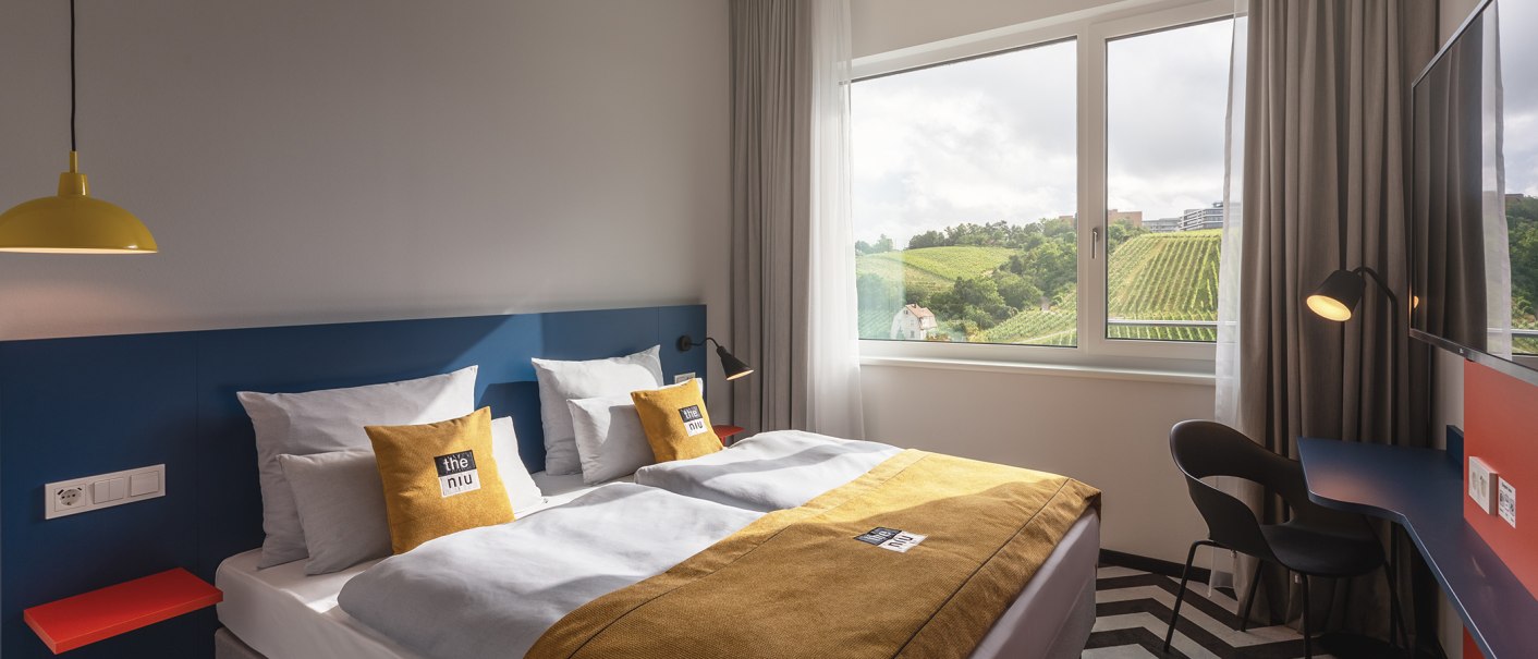 Modernes Hotelzimmer mit Doppelbett, gelben Akzenten und großem Fenster mit Blick auf Weinberge., © the niu Form - Novum Hospitality