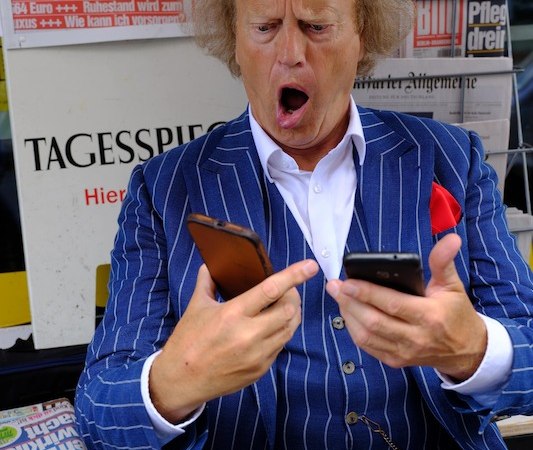 Ein Mann im gestreiften Anzug schaut überrascht auf zwei Smartphones. Im Hintergrund sind Zeitungen mit Schlagzeilen zu sehen., © Kulturhaus Schwanen