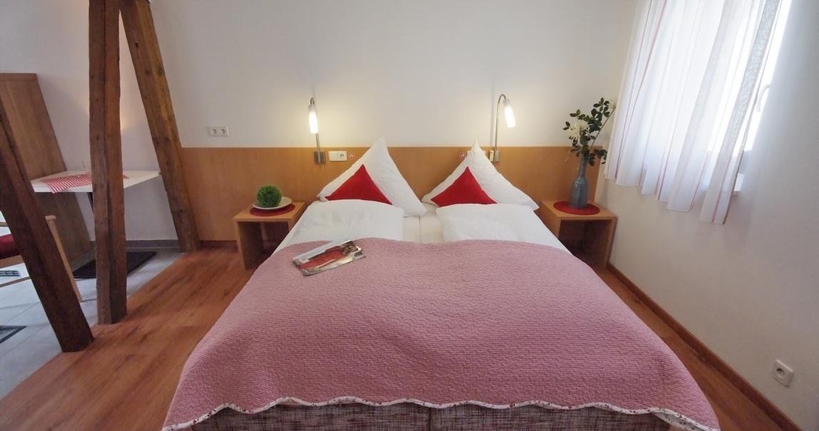 Schlafzimmer mit Doppelbett, rosa Decke, roten Kissen, Holzfußboden und Nachttischen. Helle Vorhänge und Pflanzen dekorieren den Raum., © TOMAS
