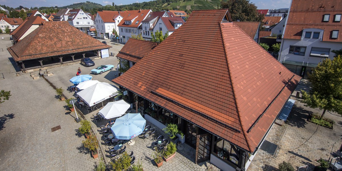 Luftaufnahme der Sieben Keltern in Metzingen. Rote Ziegeldächer, Sonnenschirme und Tische auf einem gepflasterten Platz. Umgeben von Gebäuden und grünen Hügeln., © Stuttgart-Marketing GmbH