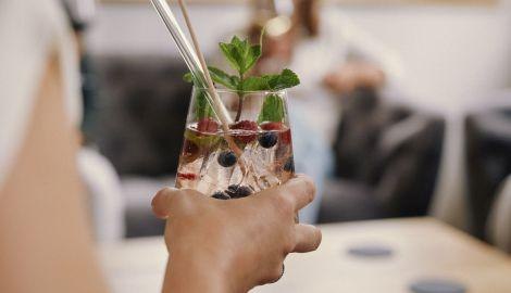Ein Cocktailglas mit Beeren und Minze wird von einer Hand gehalten. Im Hintergrund sind unscharf Personen zu erkennen., &copy; Jubil&auml;ums.Party. Foto: Teamwerk