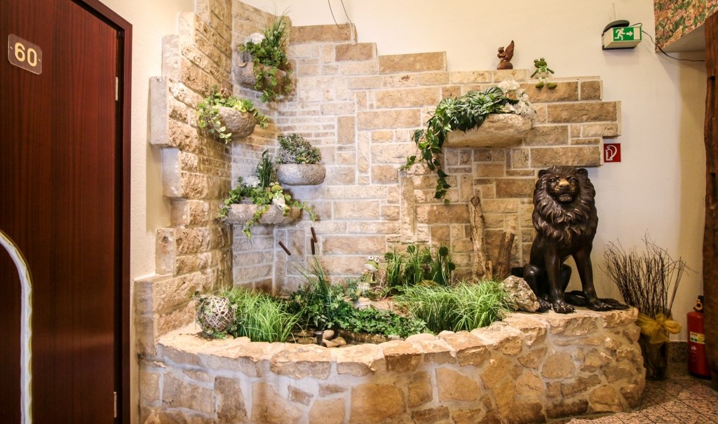 Steinwand mit Pflanzenarrangement und Löwenstatue in einer Halle. Tür mit Nummer 60 und Notausgangsschild sichtbar., © Ulli Kellenbenz Steinwand mit Pflanzenarrangement und Löwenstatue in einer Halle. Tür mit Nummer 60 und Notausgangsschild sichtbar., © Ulli Kellenbenz
