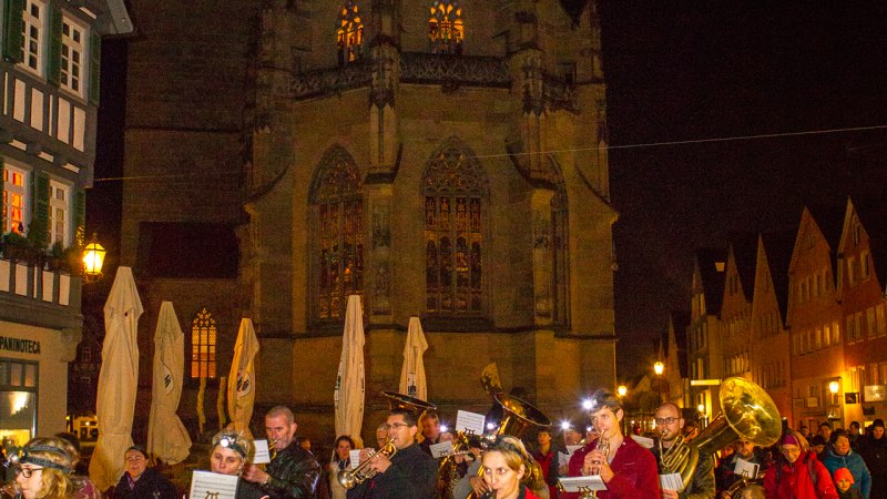 Menschen marschieren bei Nacht mit Blasinstrumenten durch Schorndorf, vorbei an einer beleuchteten Kirche und Fachwerkhäusern., © Stadt Schorndorf Menschen marschieren bei Nacht mit Blasinstrumenten durch Schorndorf, vorbei an einer beleuchteten Kirche und Fachwerkhäusern., © Stadt Schorndorf