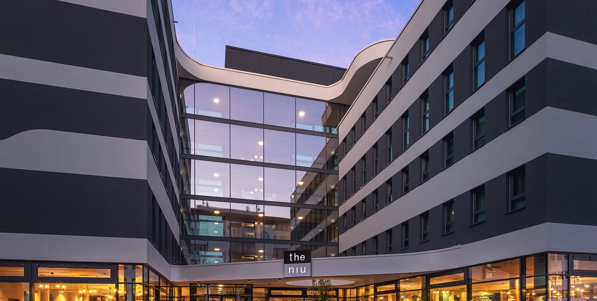 Moderne Hotelaußenfassade mit Glasfront und beleuchtetem Eingangsbereich bei Dämmerung. Das Gebäude hat ein elegantes, wellenförmiges Design., © Novum Hospitality Moderne Hotelaußenfassade mit Glasfront und beleuchtetem Eingangsbereich bei Dämmerung. Das Gebäude hat ein elegantes, wellenförmiges Design., © Novum Hospitality