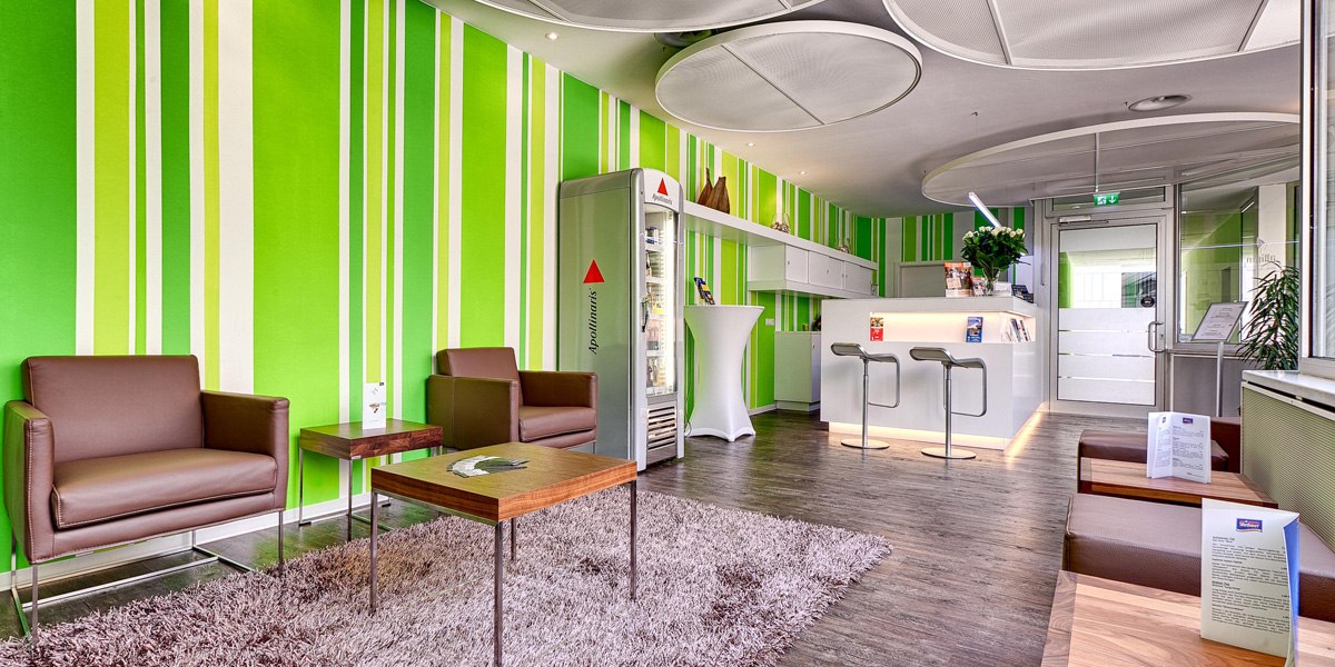 Moderne Rezeption mit grüner gestreifter Wand, braunen Sesseln, Teppich und weißem Tresen. Helle, einladende Atmosphäre mit Pflanzen und Broschüren., © attimo
