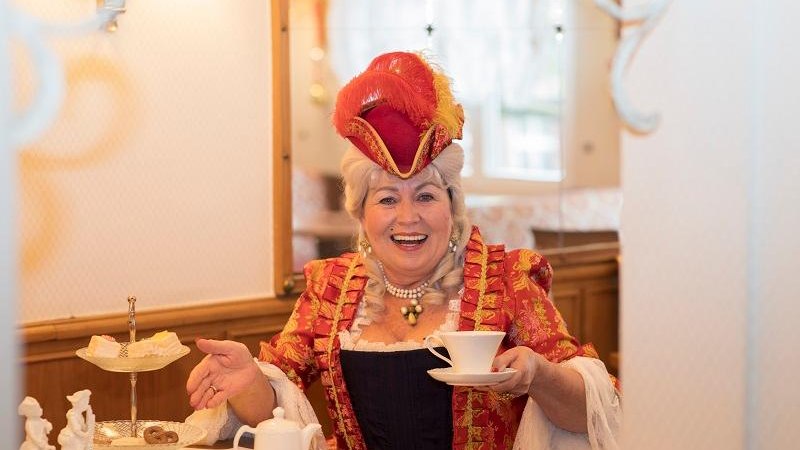 Frau in historischer Kleidung mit rotem Hut und Perlenkette genießt Kaffee in einem Café. Auf dem Tisch stehen Gebäck und eine Kaffeekanne., © Fotografin: Jasmin Schuler Frau in historischer Kleidung mit rotem Hut und Perlenkette genießt Kaffee in einem Café. Auf dem Tisch stehen Gebäck und eine Kaffeekanne., © Fotografin: Jasmin Schuler
