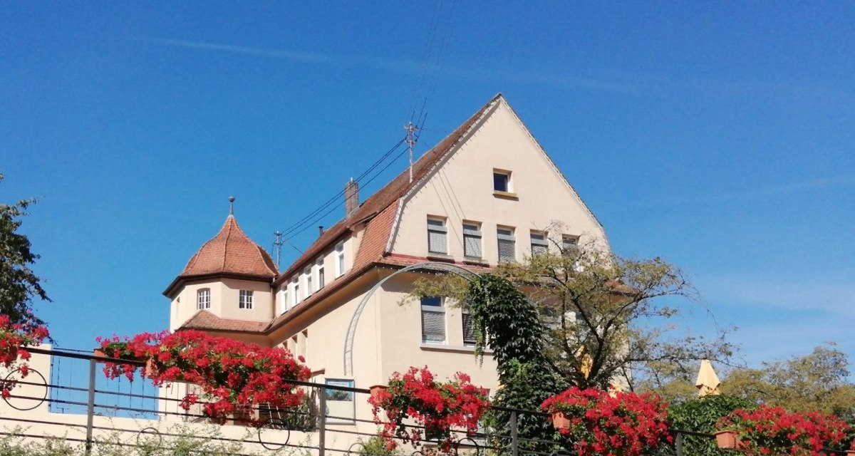Das alte Schulhaus Bargau mit roten Dachziegeln und einem Turm. Im Vordergrund blühende rote Blumen und ein klarer blauer Himmel., © Foto: Cornelia Steinbach Das alte Schulhaus Bargau mit roten Dachziegeln und einem Turm. Im Vordergrund blühende rote Blumen und ein klarer blauer Himmel., © Foto: Cornelia Steinbach