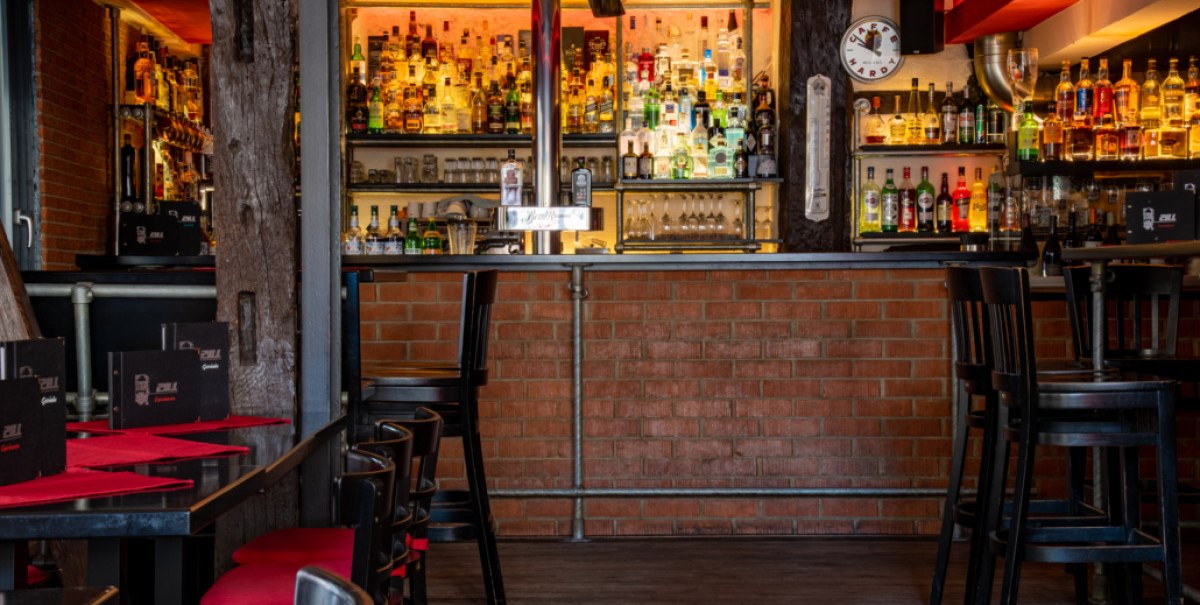 Gemütliche Bar mit Backsteinwand, gut bestücktem Regal und roten Sitzpolstern. Schwarze Tische und Stühle, warme Beleuchtung., © Café Bar Zill