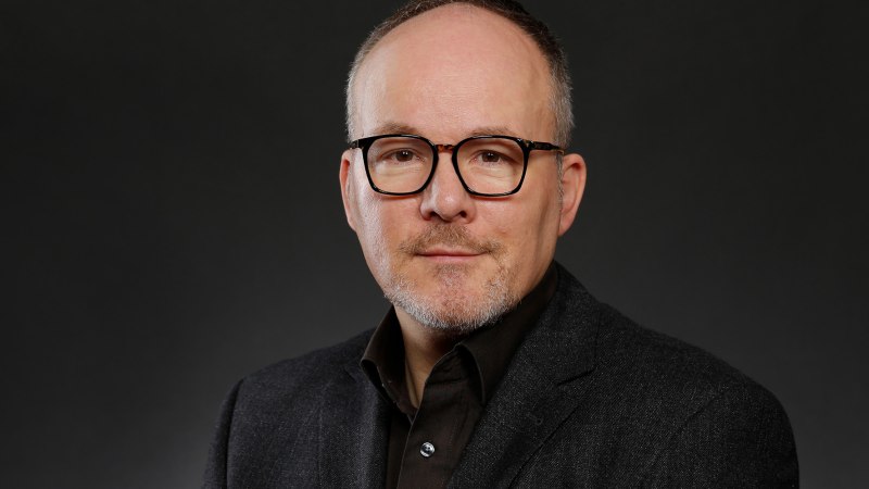 Ein Mann mit Brille und Bart in einem dunklen Anzug vor einem neutralen, dunklen Hintergrund.