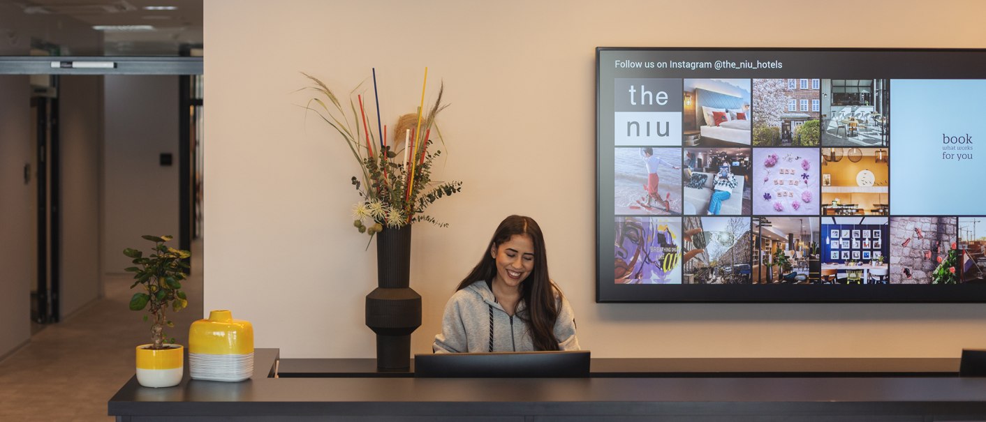 Frau an Hotelrezeption, lächelnd, mit Pflanzen und Bildschirm mit Instagram-Bildern im Hintergrund., © the niu Form - Novum Hospitality