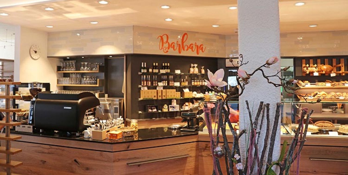 Ein gemütliches Café mit einer Theke, Kaffeemaschine und dekorativen Blumen. Im Hintergrund sind Regale mit Produkten und ein Schild mit 'Barbara' zu sehen., © Biosphärenhotel Graf Eberhard
