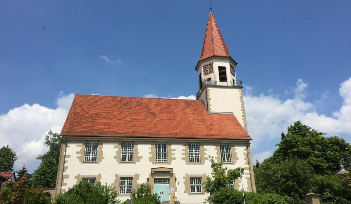 Die Kirche in Gärtringen mit rotem Dach und Turm erhebt sich vor einem klaren blauen Himmel, umgeben von grünen Bäumen und Sträuchern., © www.pro-cycl.de Die Kirche in Gärtringen mit rotem Dach und Turm erhebt sich vor einem klaren blauen Himmel, umgeben von grünen Bäumen und Sträuchern., © www.pro-cycl.de