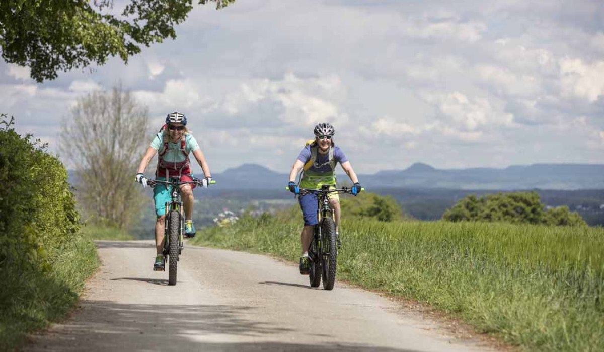 Zwei Radfahrer auf einem ländlichen Weg, umgeben von grünen Feldern und Hügeln im Hintergrund. Der Himmel ist bewölkt., © Schwäbische Alb Tourismusverband e.V.