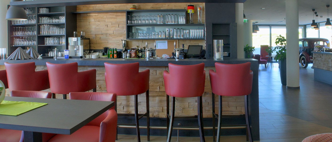 Moderne Hotelbar mit roten Barhockern, Gläsern im Regal und einem klassischen Auto im Hintergrund. Helle, einladende Atmosphäre., © TOMAS