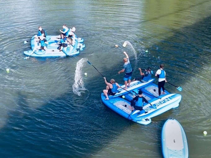 Zwei Gruppen auf blauen SUP-Boards spielen im Wasser. Einige Personen spritzen Wasser mit Paddeln, während andere zuschauen., © Cool-Tours StattReisen Zwei Gruppen auf blauen SUP-Boards spielen im Wasser. Einige Personen spritzen Wasser mit Paddeln, während andere zuschauen., © Cool-Tours StattReisen