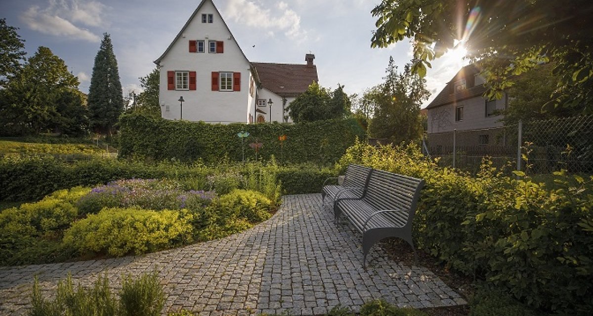Ein gepflegter Garten mit B&auml;nken, gepflastertem Weg und einem wei&szlig;en Haus im Hintergrund. Die Sonne scheint durch die B&auml;ume., &copy; Stoppel