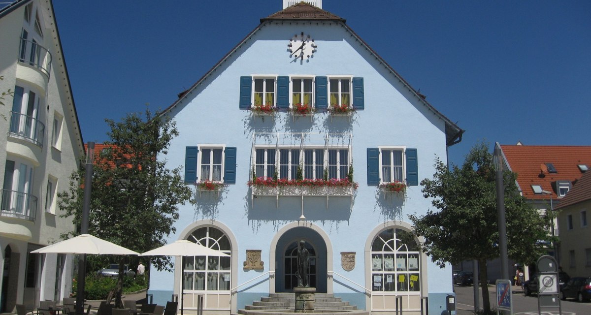 Blaues Gebäude mit Uhr und Blumenkästen, davor ein Brunnen und Café mit Sonnenschirmen, bei sonnigem Wetter., © http://www.rutesheim.de/servlet/PB/menu/1110073/index.html