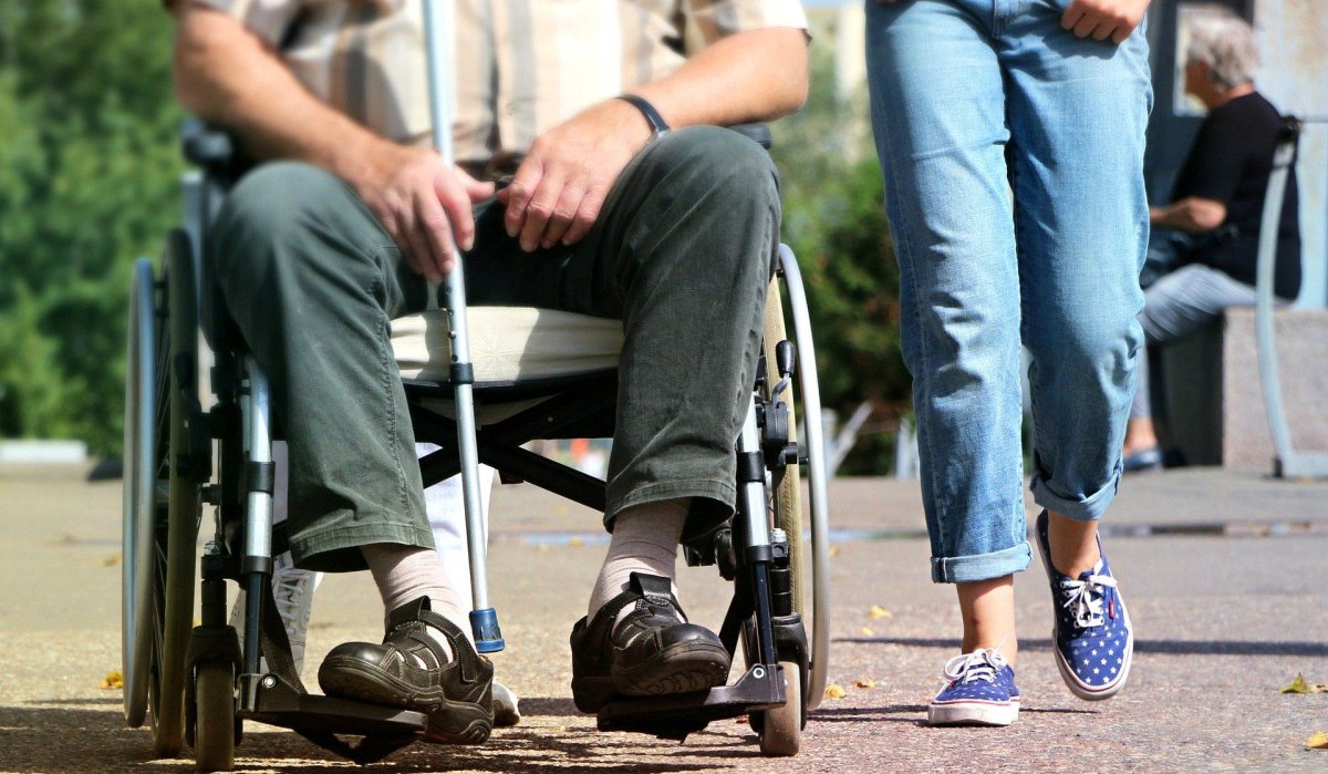 Ein Mann sitzt in einem Rollstuhl auf einem Gehweg, während eine Person in Jeans und Turnschuhen daneben geht., © pixabay Ein Mann sitzt in einem Rollstuhl auf einem Gehweg, während eine Person in Jeans und Turnschuhen daneben geht., © pixabay