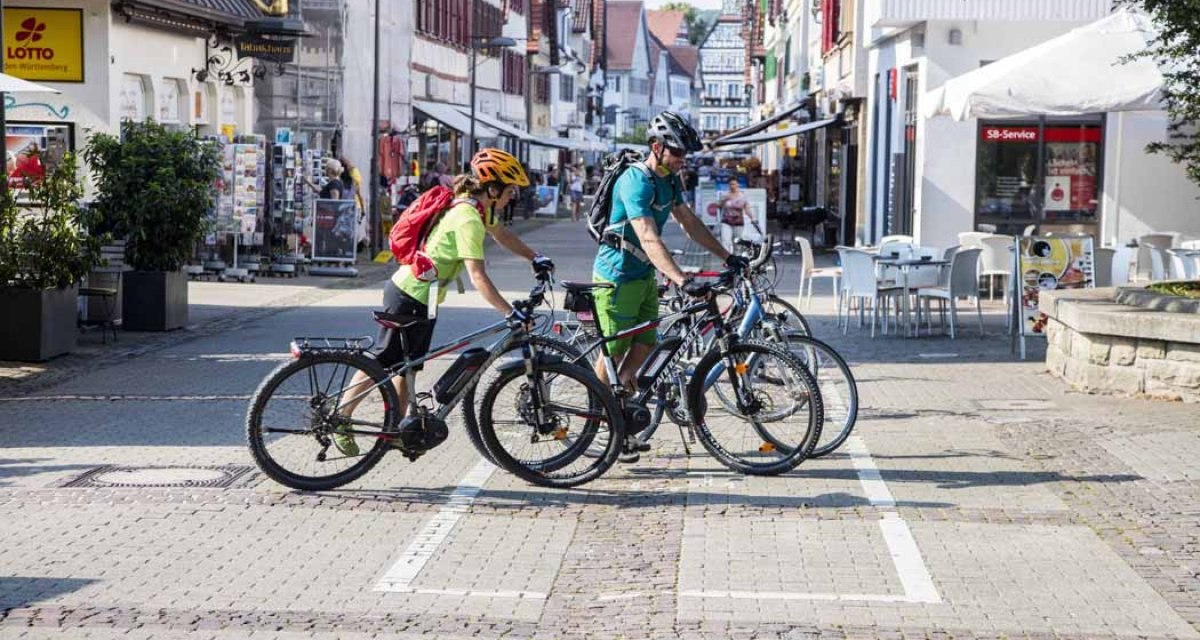 Zwei Radfahrer schieben ihre Fahrräder durch eine belebte Fußgängerzone in Kirchheim unter Teck. Im Hintergrund sind Geschäfte und Cafés zu sehen., © Schwäbische Alb Tourismusverband e.V. Zwei Radfahrer schieben ihre Fahrräder durch eine belebte Fußgängerzone in Kirchheim unter Teck. Im Hintergrund sind Geschäfte und Cafés zu sehen., © Schwäbische Alb Tourismusverband e.V.