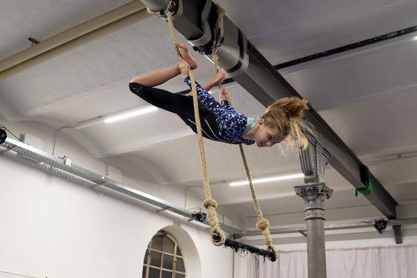 Ein Mädchen in einem blauen Anzug macht Luftakrobatik an Seilen in einer Halle. Sie hängt kopfüber und zeigt eine beeindruckende Pose., © Kunstschule Labyrinth