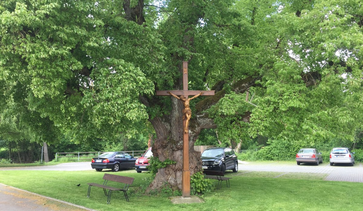 Holzkreuz mit Jesusfigur vor einem großen Baum, umgeben von geparkten Autos und einer Bank auf einer grünen Wiese., © www.pro-cycl.de Holzkreuz mit Jesusfigur vor einem großen Baum, umgeben von geparkten Autos und einer Bank auf einer grünen Wiese., © www.pro-cycl.de