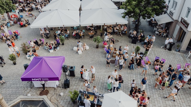 Belebter Platz in Metzingen mit Menschen unter wei&szlig;en Sonnenschirmen und einem lila Zelt. Viele Besucher genie&szlig;en die entspannte Atmosph&auml;re.