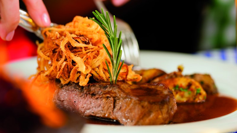 Zwiebelrostbraten mit knusprigen Röstzwiebeln und Rosmarin auf einem Teller, im Hintergrund unscharf gedeckter Tisch., © Fotolia, Kzenon