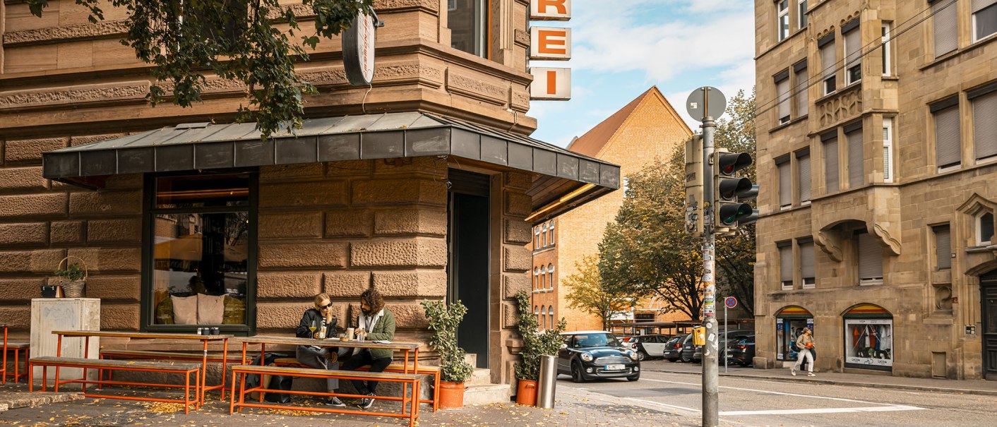 Eine Straßenecke mit einer Metzgerei. Zwei Personen sitzen an einem Tisch vor dem Laden. Ein Auto fährt vorbei, und es gibt eine Ampel., © SMG, Sarah Schmid