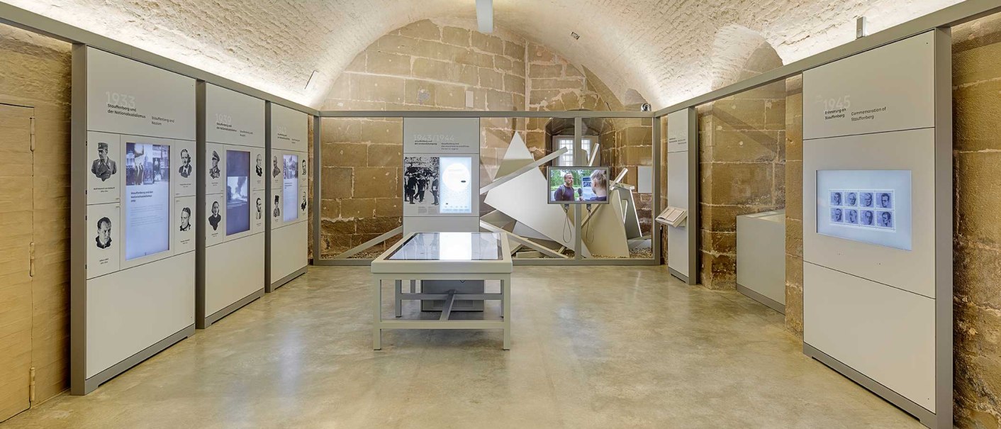 Innenansicht der Stauffenberg-Erinnerungsstätte mit modernen Ausstellungswänden, interaktiven Displays und einem zentralen Tisch in einem Gewölberaum., © Haus der Geschichte Baden-Württemberg Innenansicht der Stauffenberg-Erinnerungsstätte mit modernen Ausstellungswänden, interaktiven Displays und einem zentralen Tisch in einem Gewölberaum., © Haus der Geschichte Baden-Württemberg