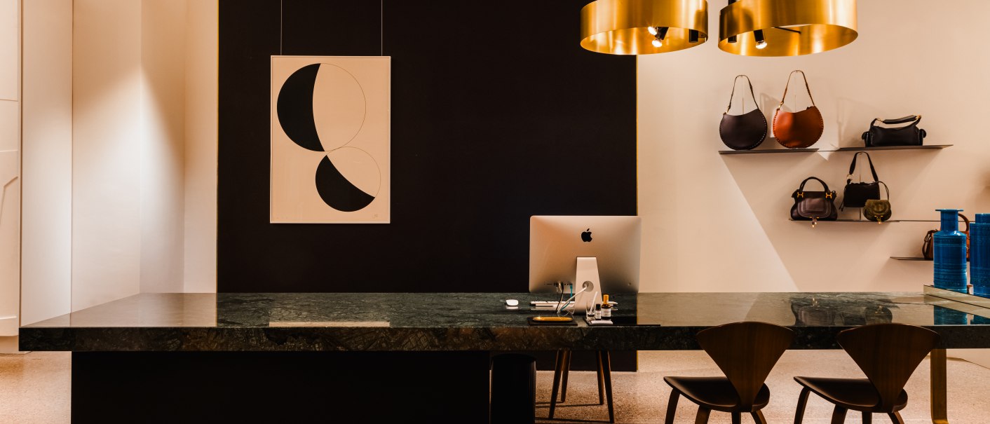 Eleganter Laden mit großem Tisch, zwei Stühlen, Computer und Regalen mit Handtaschen. Moderne Beleuchtung und abstrakte Kunst an der Wand., © Bungalow