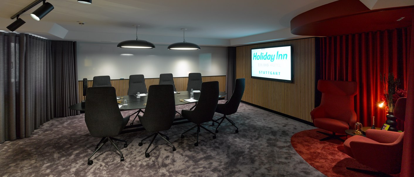 Moderner Konferenzraum mit großem Tisch, schwarzen Stühlen und rotem Sitzbereich. An der Wand ein Holiday Inn Stuttgart Schild., © Holiday Inn Stuttgart Moderner Konferenzraum mit großem Tisch, schwarzen Stühlen und rotem Sitzbereich. An der Wand ein Holiday Inn Stuttgart Schild., © Holiday Inn Stuttgart