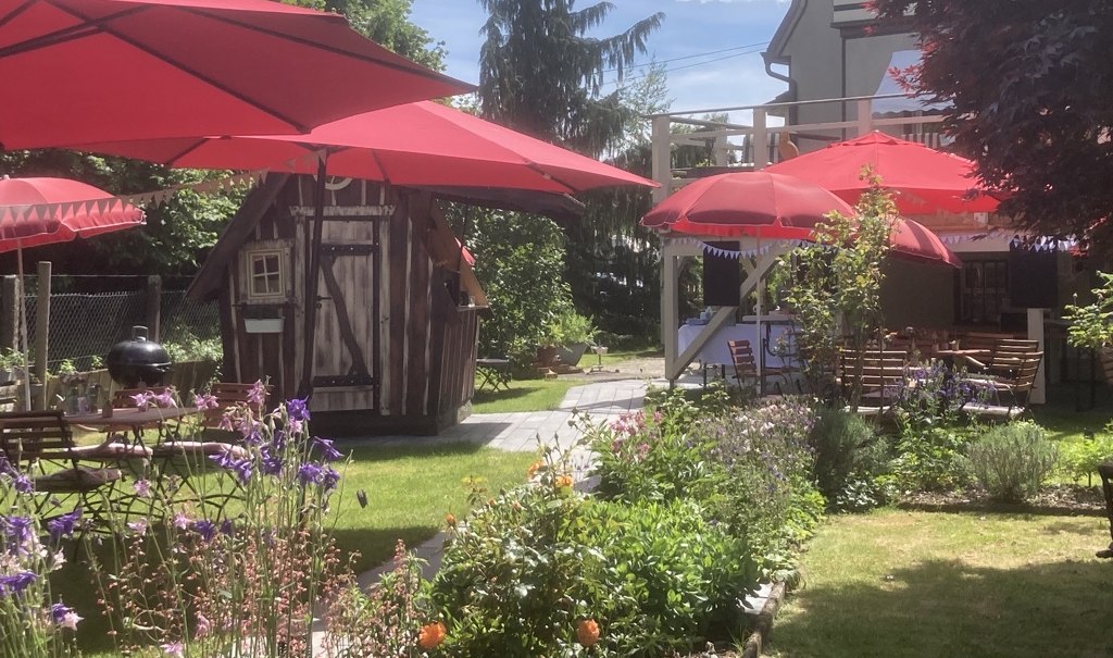 Idyllischer Garten mit roten Sonnenschirmen, Holztischen und blühenden Blumen. Ein kleines Holzhaus und ein größeres Gebäude im Hintergrund., © Café Plätschwiesle