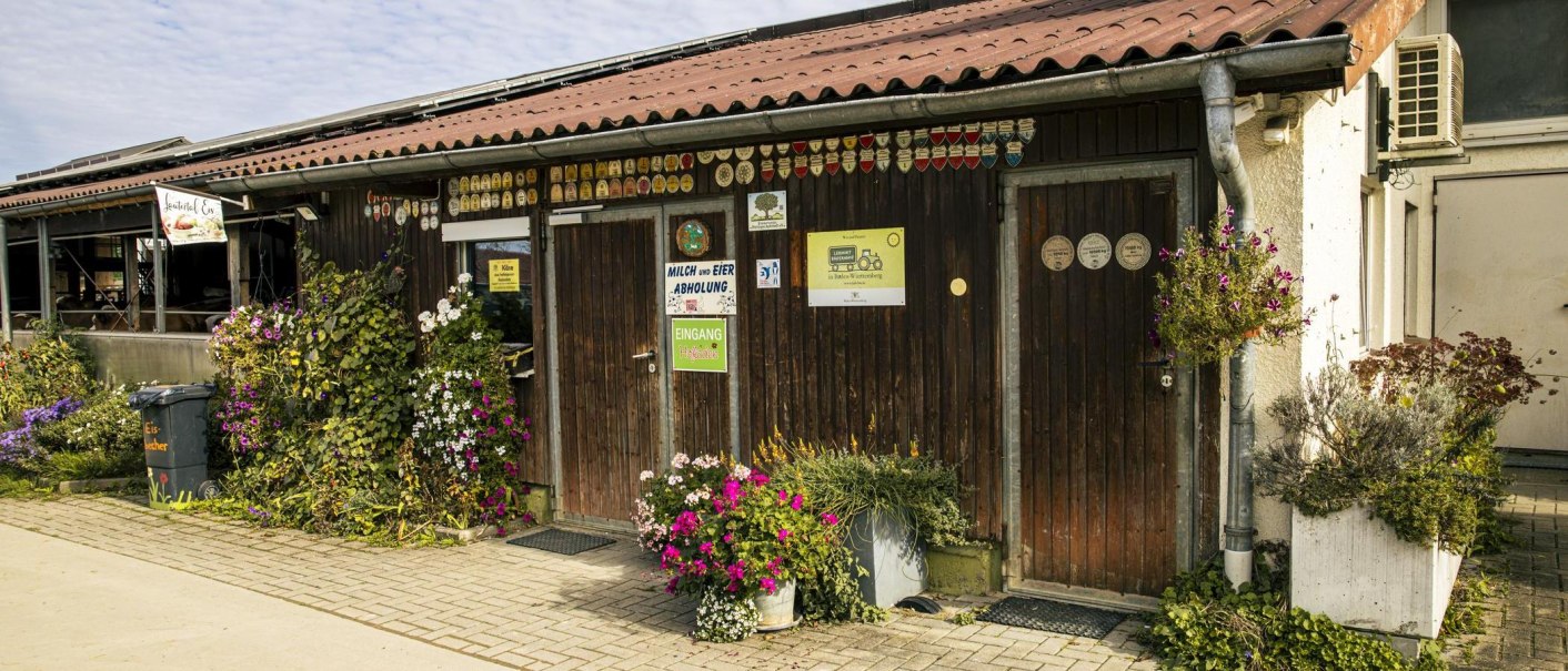 Rustikaler Hofladen mit Holzwänden, Schildern und blühenden Pflanzen. Ein Schild weist auf Milch- und Eierabholung hin. Gepflasterter Weg davor., © Stuttgart Marketing GmbH, Sarah Schmid