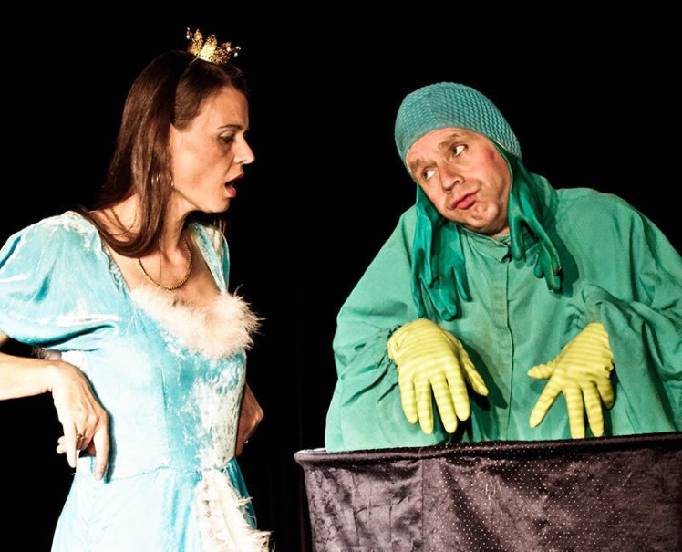Eine Frau in einem blauen Kleid mit Krone steht auf einer B&uuml;hne und spricht mit einem Mann in einem Froschkost&uuml;m. Beide sind in einem Theaterst&uuml;ck., &copy; Galli Theater Backnang