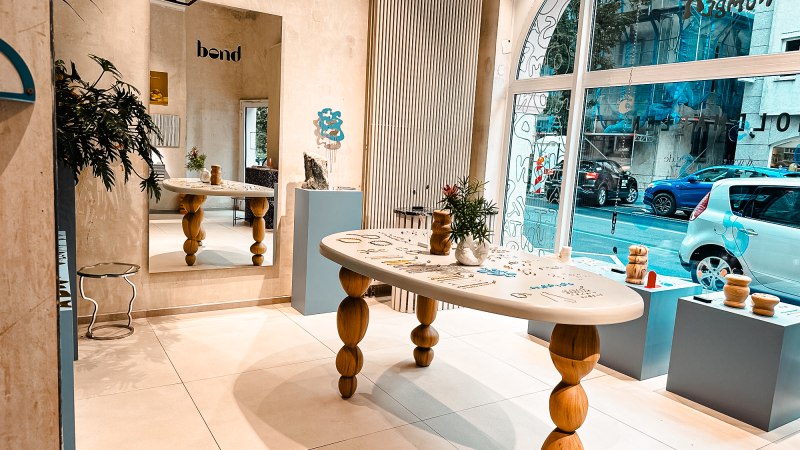 Modernes Studio mit Holztisch, Spiegel und Dekorationen. Große Fensterfront mit Blick auf die Straße. Pflanzen und Kunstobjekte als Dekoration., © Goldmarlen