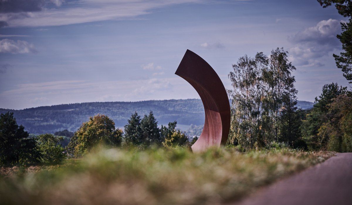 Große, gebogene Skulptur in einer Landschaft mit Bäumen und Hügeln im Hintergrund unter blauem Himmel., © Natur.Nah. Schönbuch & Heckengäu Große, gebogene Skulptur in einer Landschaft mit Bäumen und Hügeln im Hintergrund unter blauem Himmel., © Natur.Nah. Schönbuch & Heckengäu