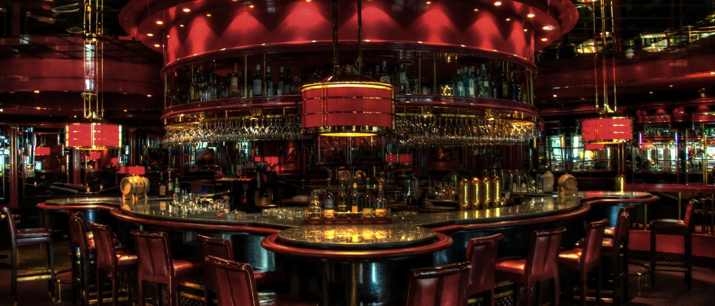 Elegante Bar mit roten und goldenen Akzenten, umgeben von Barhockern und Flaschen im Hintergrund., © Maritim Hotelgesellschaft mbH