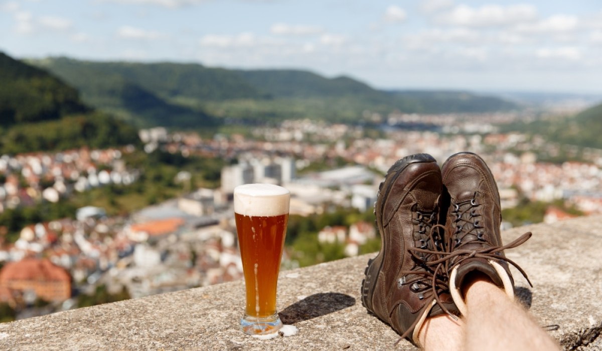 Wanderschuhe ruhen auf einer Mauer neben einem Glas Bier. Im Hintergrund ist die Stadt Geislingen und eine hügelige Landschaft zu sehen., © Landkreis Göppingen