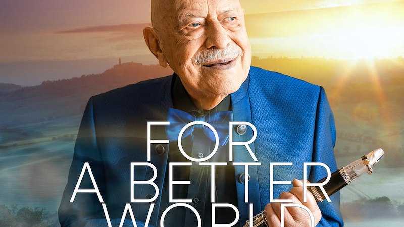 Ein &auml;lterer Mann in einem blauen Anzug h&auml;lt eine Klarinette vor einer malerischen Landschaft bei Sonnenuntergang. Text: Maestro Giora Feidman - For a Better World., &copy; MACC Management GmbH