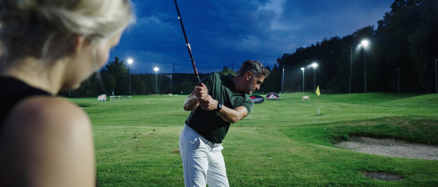 Ein Mann beim Golfschwung auf einem beleuchteten Platz in der Dämmerung, beobachtet von einer Frau im Vordergrund., © GolfKultur Stuttgart Ein Mann beim Golfschwung auf einem beleuchteten Platz in der Dämmerung, beobachtet von einer Frau im Vordergrund., © GolfKultur Stuttgart